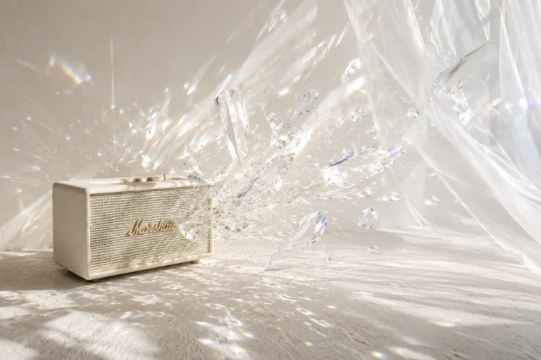 White vintage speaker blasting clear crystal and glass shards across a sunlit white room with billowing sheer curtains.