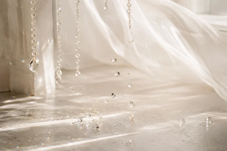 Broken strands of clear crystal beads falling and rolling across a sunlit whitewashed floor beside a billowing sheer curtain.