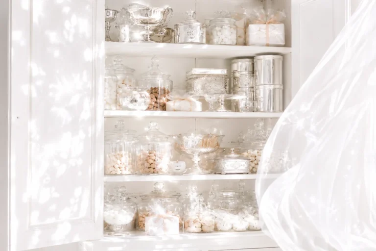 An open white vintage cupboard filled with crystal jars and silver tins, lit by dappled sunlight beside a billowing sheer curtain.
