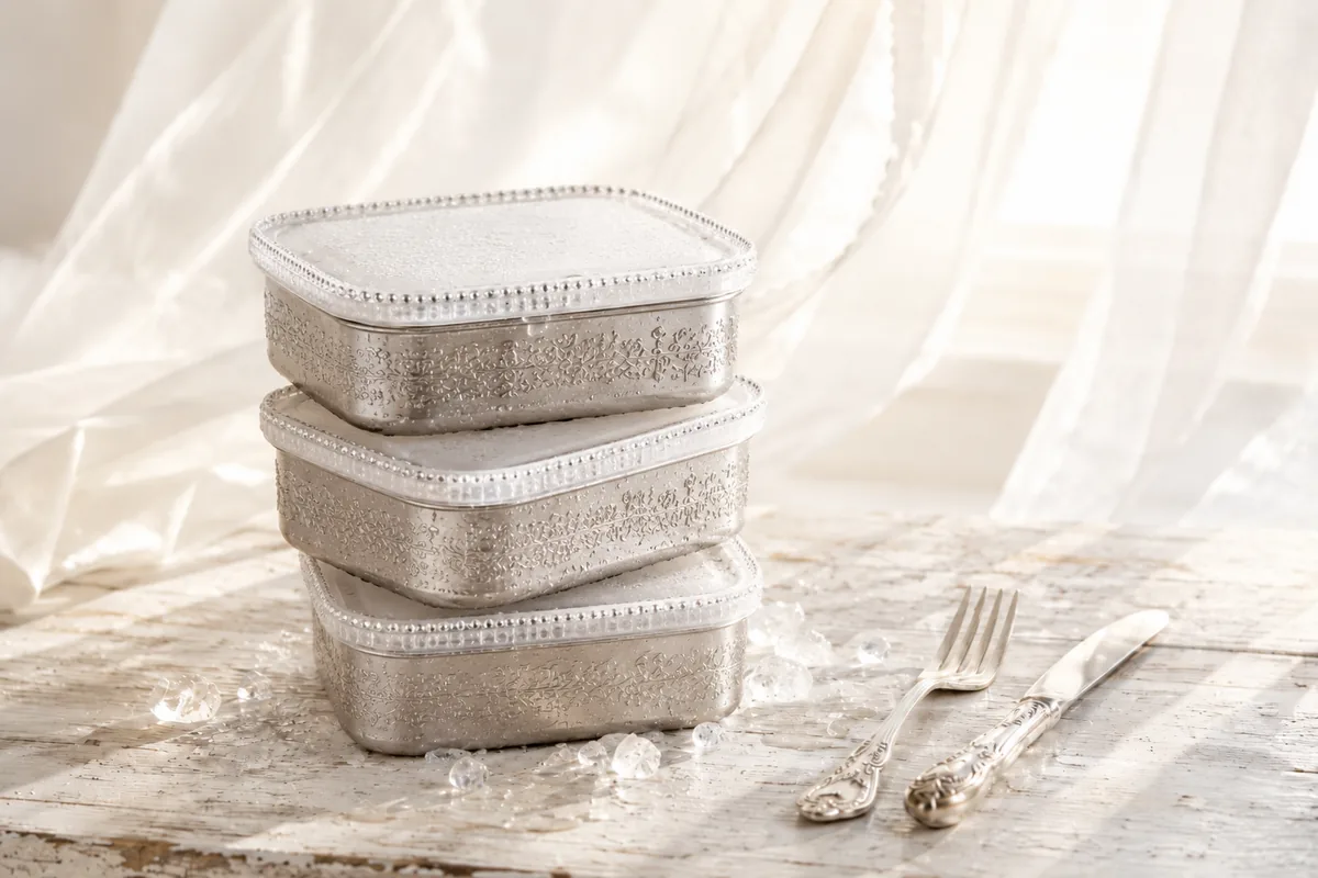 Three engraved silver freezer containers with frosted crystal-edged lids, stacked on a whitewashed table beside vintage cutlery and scattered ice.