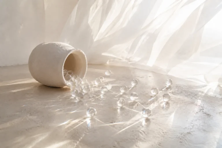 Clear glass marbles spilling from a tipped white clay jar onto a sunlit concrete floor, with a billowing white curtain behind.