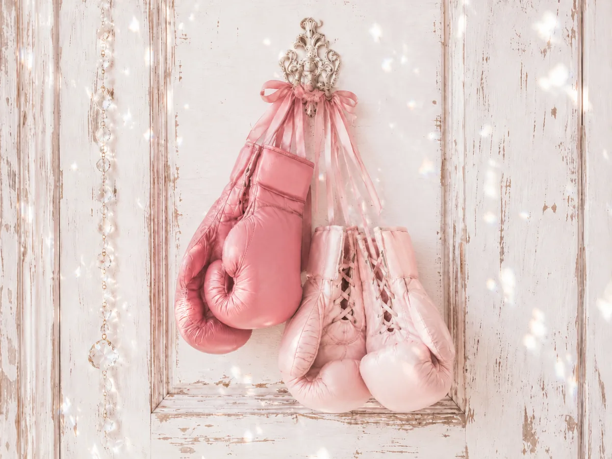 Two pairs of blush-pink boxing gloves hanging unevenly from one ornate hook on a distressed white door with crystal sparkles.