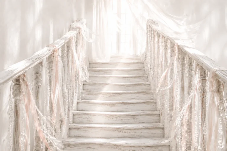 Old whitewashed wooden staircase with blush-pink ribbons and crystal strands leading towards a bright, hazy window.