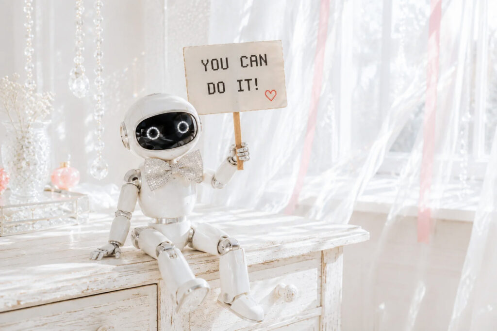Small white robot holding a “YOU CAN DO IT!” sign with a red heart on a sunlit whitewashed desk.