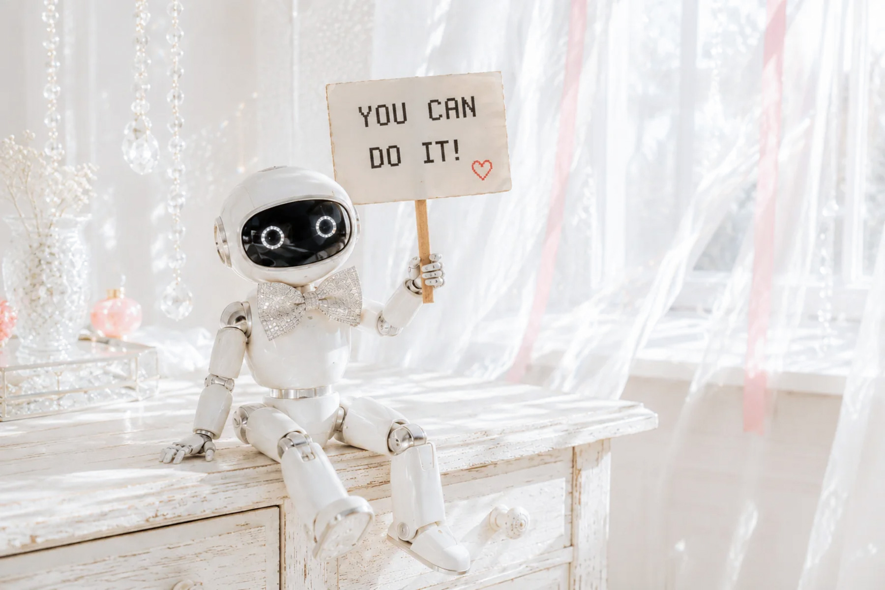 Small white robot holding a “YOU CAN DO IT!” sign with a red heart on a sunlit whitewashed desk.