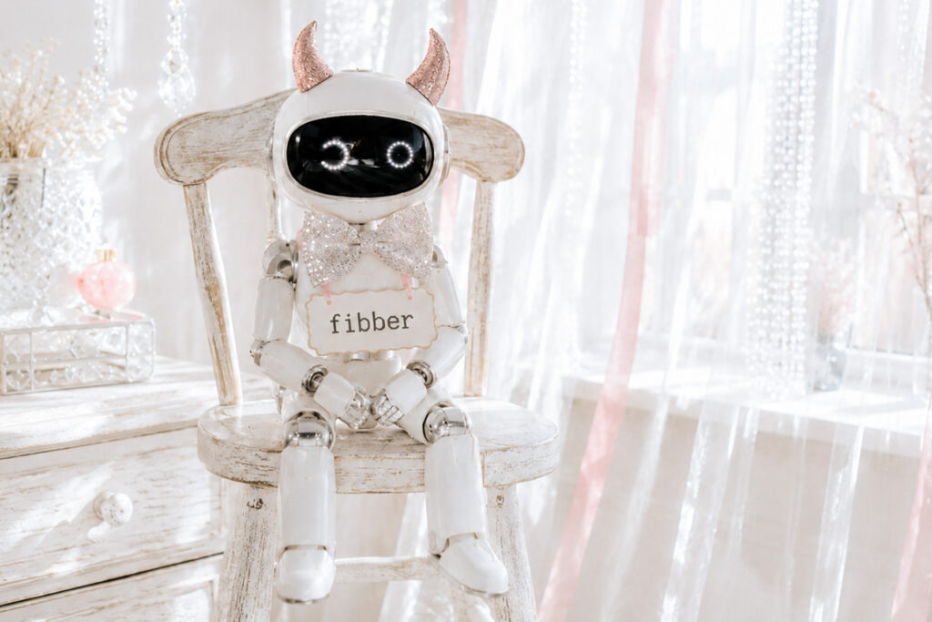 Small white robot with glittery pink devil horns sitting on a distressed white chair, wearing a sign that says “fibber” in a bright white room with sheer curtains and crystal strands.