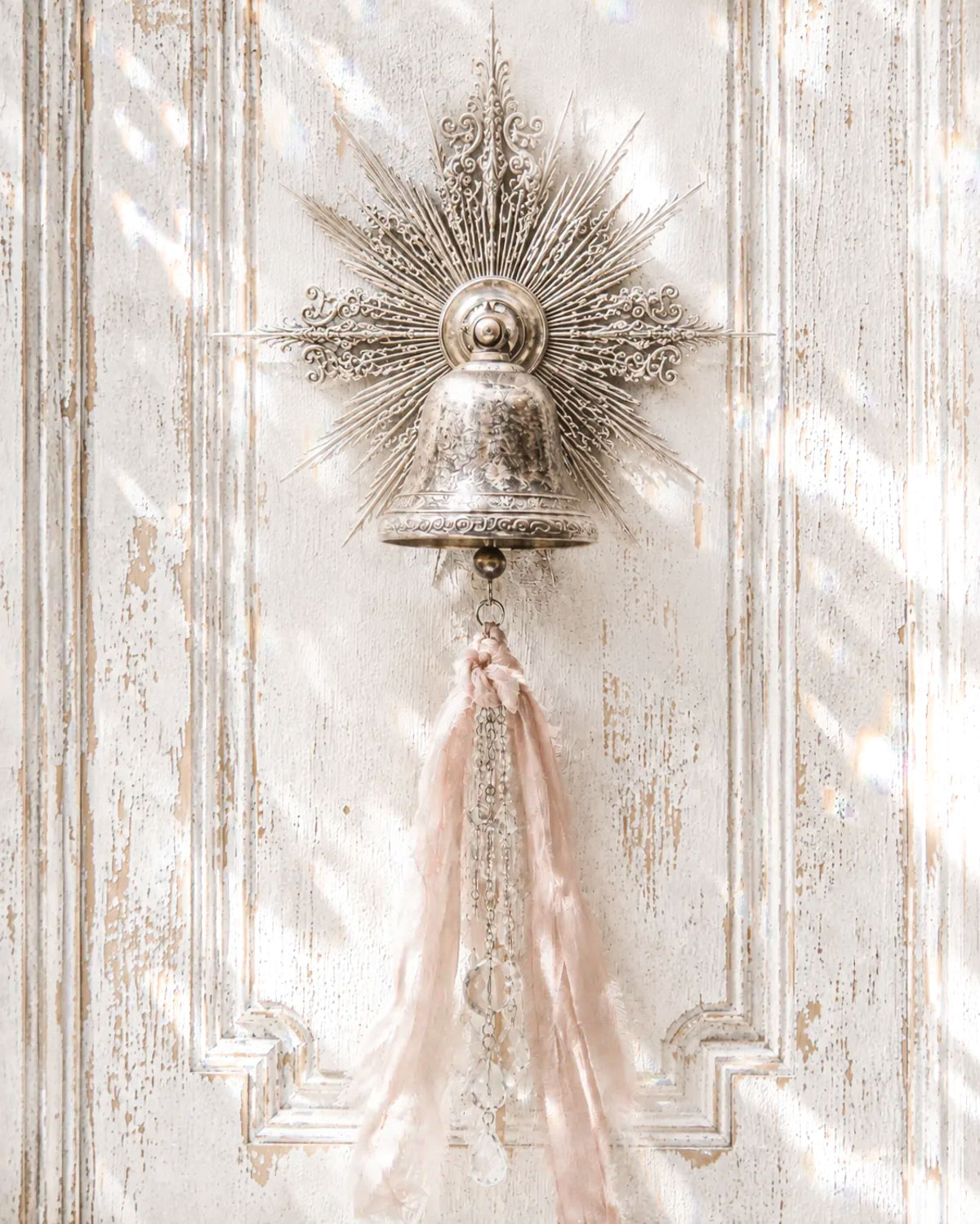 Antique silver bell with a long blush-pink ribbon and crystal pull mounted on a distressed white panelled door in bright dappled light.