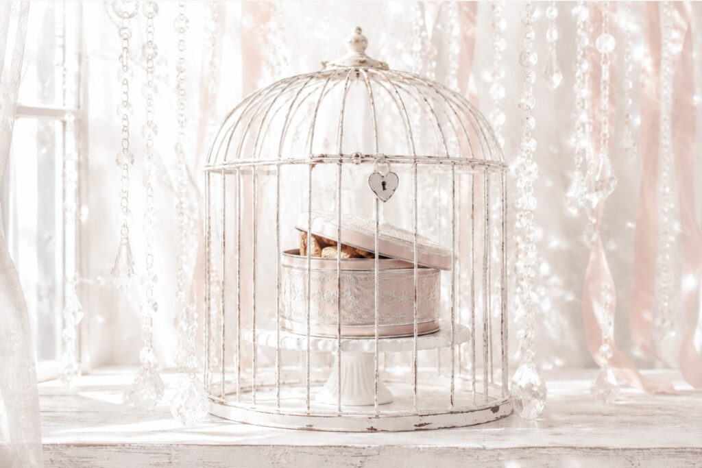 Vintage blush pink cookie tin inside a locked white birdcage on a cake stand, with soft ribbons, crystals and sunlight behind.