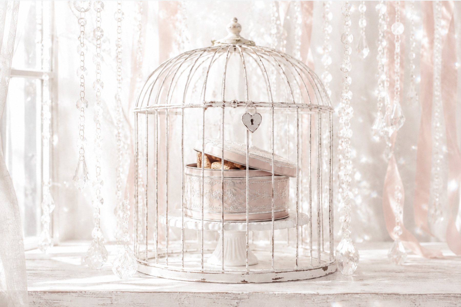 Vintage blush pink cookie tin inside a locked white birdcage on a cake stand, with soft ribbons, crystals and sunlight behind.